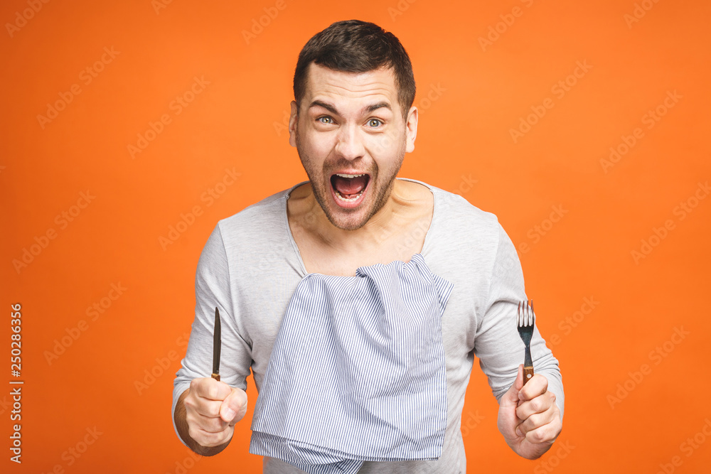 Wall mural Young hungry crazy man holding a fork and a knife. Isolated on orange background.