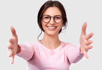 Closeup shot of happy pretty brunette young woman shows welcome gesture, spreads hands as wants to cuddle someone, smiles broadly, wears pink sweater and eyeglasses, isolated over white background