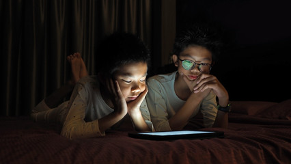 Asian brothers lying in bed at night and playing digital tablet together.