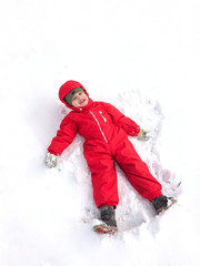 Cute little boy in a red winter jumpsuit in the winter on the street makes a snow angel and have fun.