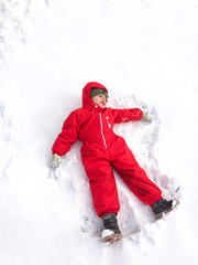 Fototapeta na wymiar Cute little boy in a red winter jumpsuit in the winter on the street makes a snow angel and have fun.