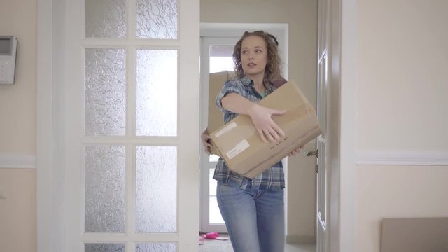 The door opens, tall man and pretty woman in casual dress with boxes in hands enters half empty room. Young people put boxes on the floor and look around Young people moving to new apartment.