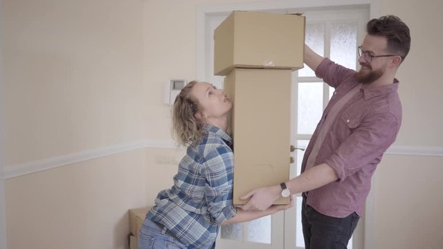 Tall man standing in the room with big box in hands. Woman takes the boxes from her husband and balances them in their arms. Married couple moves into a new home