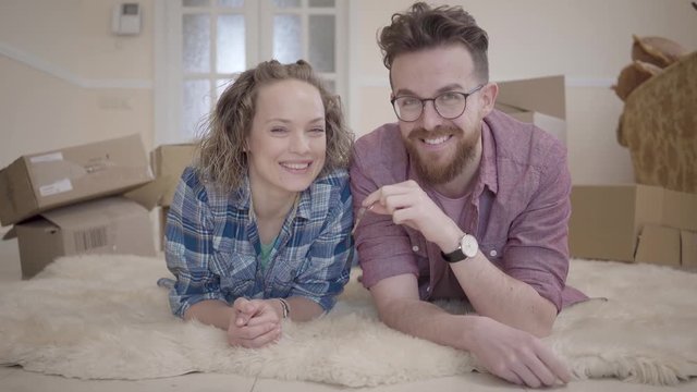 Young family lying on fluffy carpet close up with boxes in the background. Man holding a key and shakes it, looking in camera. Married couple moves into a new home
