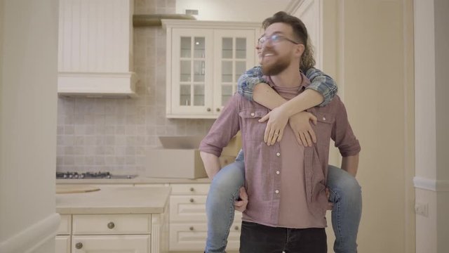 Bearded man carries pretty woman sitting on his back in the kitchen and puts her on the table. Woman hugs her husband, they kissing. Married couple moves into a new home