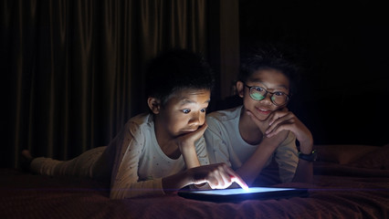 Asian brothers lying in bed at night and playing digital tablet together with surprise faces.