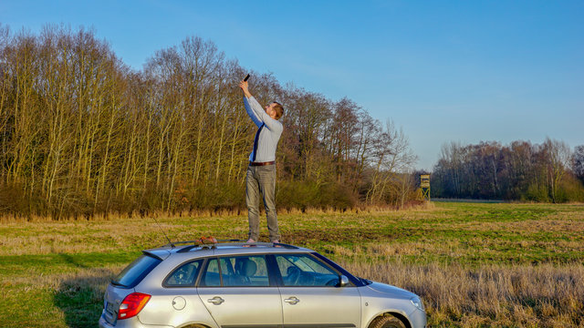 Mann steht auf einem Autodach und versucht Empfang zu finden, um zu telefonieren, weil er eine Autopanne hat