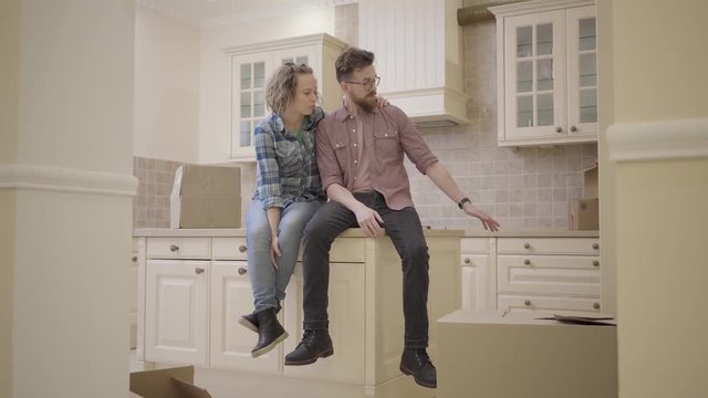 Bearded man and pretty woman sitting on the table in kitchen of new apartment. Family discuss plans about new flat. Married couple moves into a new home. Camera moves to the right