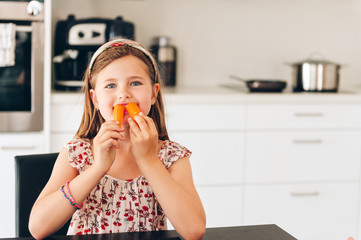 Cute little girl eating fresh carrot in white kitchen, vitamins nutrition for children