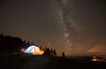 Night camping in mountains near forest. Bach view couple hikers covered by blanket plaid resting together, sitting beside campfire and tourist tent under night starry sky full of stars and Milky way.