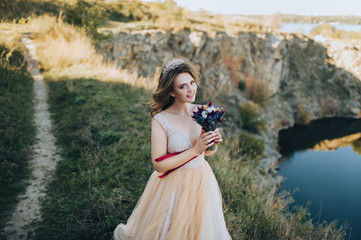 Fototapeta na wymiar Portrait of a beautiful bride in a beige dress with a crown on her head. A lovely bride with a bouquet is standing on a cliff in autumn weather. Wedding photography. Beautiful landscape.