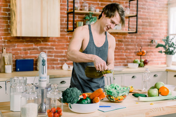 A sporty man is preparing a salad in the kitchen. He pours olive oil into a salad jar. He is standing in a modern kitchen at home.