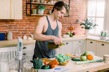 A sporty man is preparing a salad in the kitchen. He pours olive oil into a salad jar. He is standing in a modern kitchen at home.