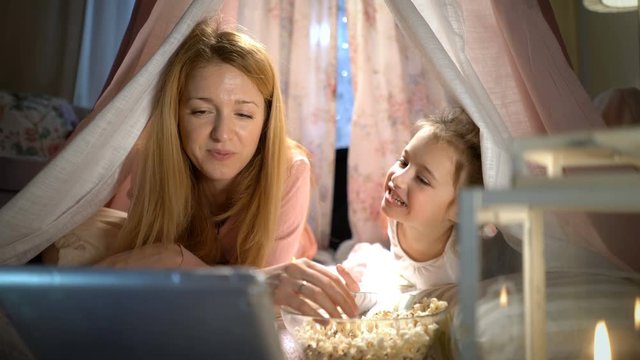 Family movie theater. Little girl and her mother enjoying watching cartoons online and eating popcorn in the tent in the nursery