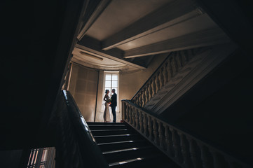 Obraz na płótnie Canvas Newlyweds in love stand in an old dark building near the bright window and embrace. Portrait of a stylish groom with glasses and a beautiful cute bride in a white dress. Wedding photography.