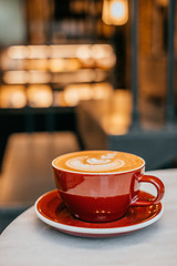 Red cup of cappuccino with beautiful latte art on round white marble table and blurred lights background.