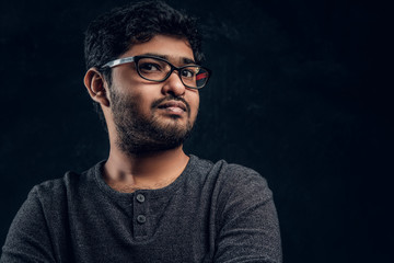 Close-up portrait of a young Indian guy in eyewear and casual clothes looking at a camera in studio against the background of the dark wall.