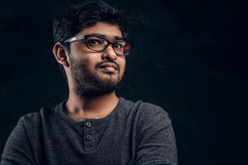 Obraz premium Close-up portrait of a young Indian guy in eyewear and casual clothes looking at a camera in studio against the background of the dark wall.