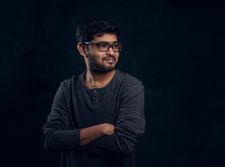 Studio portrait of a young Indian guy wearing eyewear and casual clothes posing with his arms crossed and looking sideways in a dark room