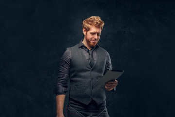 Handsome redhead man dressed in an elegant vest with tie and kilt using a tablet in studio against a dark textured wall