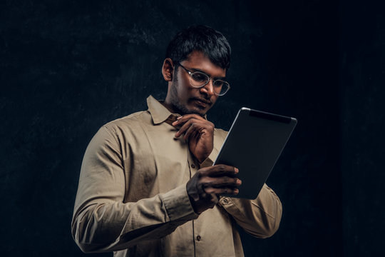 Pensive Indian Male In Eyewear And Shirt Using A Tablet Computer In Studio Against The Background Of The Dark Wall