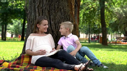 Naklejka na ściany i meble Boy runs to pregnant mother reading book in park, healthy and happy pregnancy