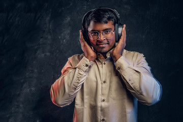Handsome Indian man enjoying listening to music in wireless headphones standing in studio against the background of the dark wall