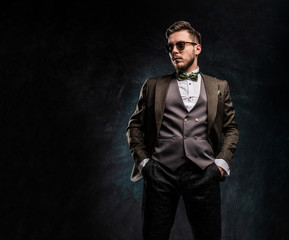 A stylish young man in sunglasses dressed in an elegant suit posing with a hand in a pocket against a dark textured wall