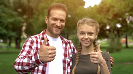 Cheerful father and teenage daughter smiling into camera, showing thumbs up