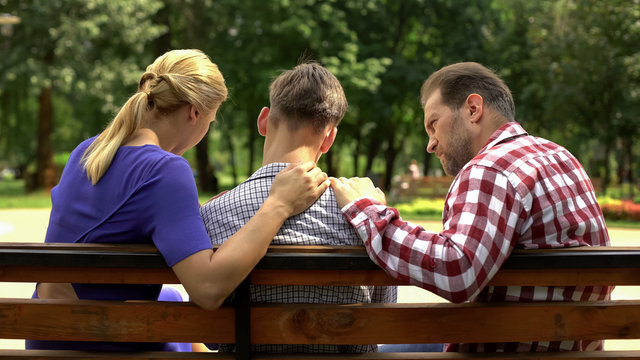 Caring mother and dad supporting sad teen son sitting on bench in park, crisis