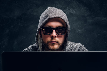 Close-up portrait of a hacker in hoodie and sunglasses sits behind laptop in office against a dark...