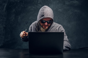 Bearded hacker in the hood looking at the laptop holding up his hand and smiling on a black...