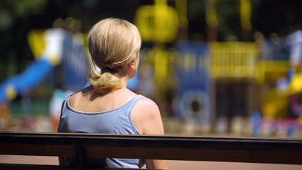 Sad old female sitting on playground bench and looking at children, infertility