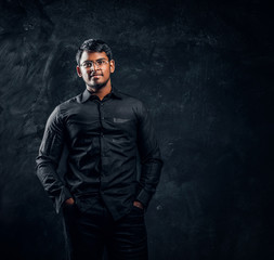 Young confident Indian guy in glasses and black shirt posing for camera holding hands in pockets. Studio photo