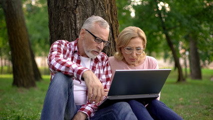 Fototapeta premium Old man and woman sitting on grass and watching thriller movie trailer on laptop