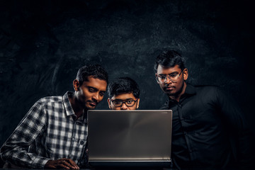 Three Indian students working on a project together standing at the table with a laptop in a dark room