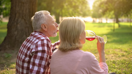 Cheerful couple of pensioners enjoying romantic date in park and drinking wine
