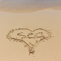 The word Sea is written inside a drawing heart on the sand on the beach against the background of a sea wave. Background for a travel, beach holiday, spa resorts, cruises with copy space