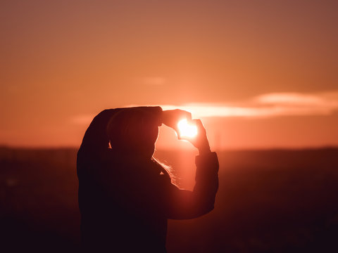 Image of woman from behind forming a heart with her hands with sun shining towards her face