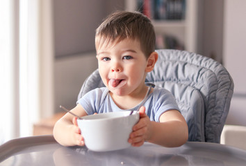 Adorable thankful little baby boy with smeared face and tongue out just finished tasty meal and hold white bowl looking at camera wanting more sitting at high feeding chair. Child nutrition concept