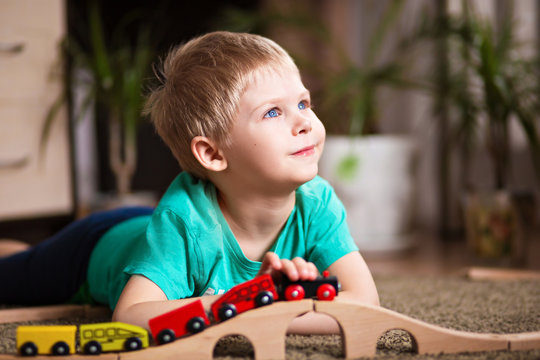 happy baby, blonde with blue eyes, a little boy of five years in the mint (green / blue) t-shirt in shorts barefoot near the couch in the apartment igret fun with wooden railway
