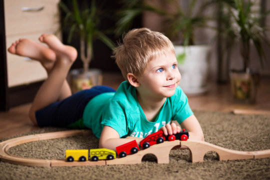 happy baby, blonde with blue eyes, a little boy of five years in the mint (green / blue) t-shirt in shorts barefoot near the couch in the apartment igret fun with wooden railway