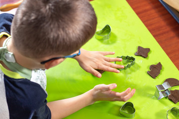 Chłopiec formuje i rozgniata brązowe ciasto dłonią. Gotowe ciastka oraz drewniany wałek leżą obok na blacie. © Tomasz