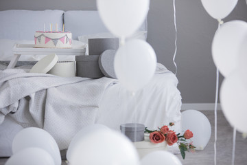 Birthday cake with candles on wooden tray next to white and grey round boxes on comfortable king size bed