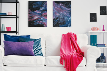 Purple, blue and green pillows and pink blanket on white sofa in stylish living room with galaxy graphic on the wall