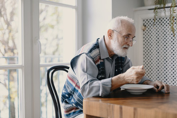 Senior grandfather with grey hair and beard sitting alone in the kitchen eating breakfast