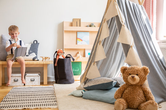 Teddy bear next to grey scandinavian tent in stylish boy's bedroom with furniture made from natural materials, real photo with copy space on the white wall