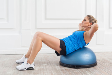 Focused sporty beautiful young athletic blonde woman in black shorts and blue top working in gym doing exercises for abdominal muscles on bosu balance trainer, lie down on fitness ball, indoor