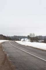 Rural asphalt road in winter. Dirty road with cracks. Winter fields and forest on a cloudy day