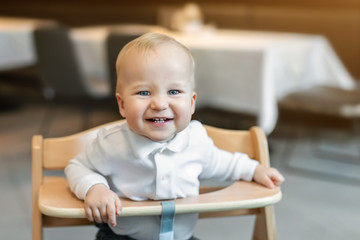 Cute little baby boy in white polo t-shirt sitting in wooden baby chair and laughing at cafe indoors. Portrait of adorable blond kid having fun at restaurant